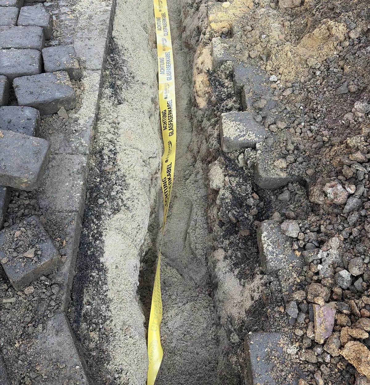 Schmaler Kabelgraben im aufgenommenen Pflaster mit gelbem Warnband