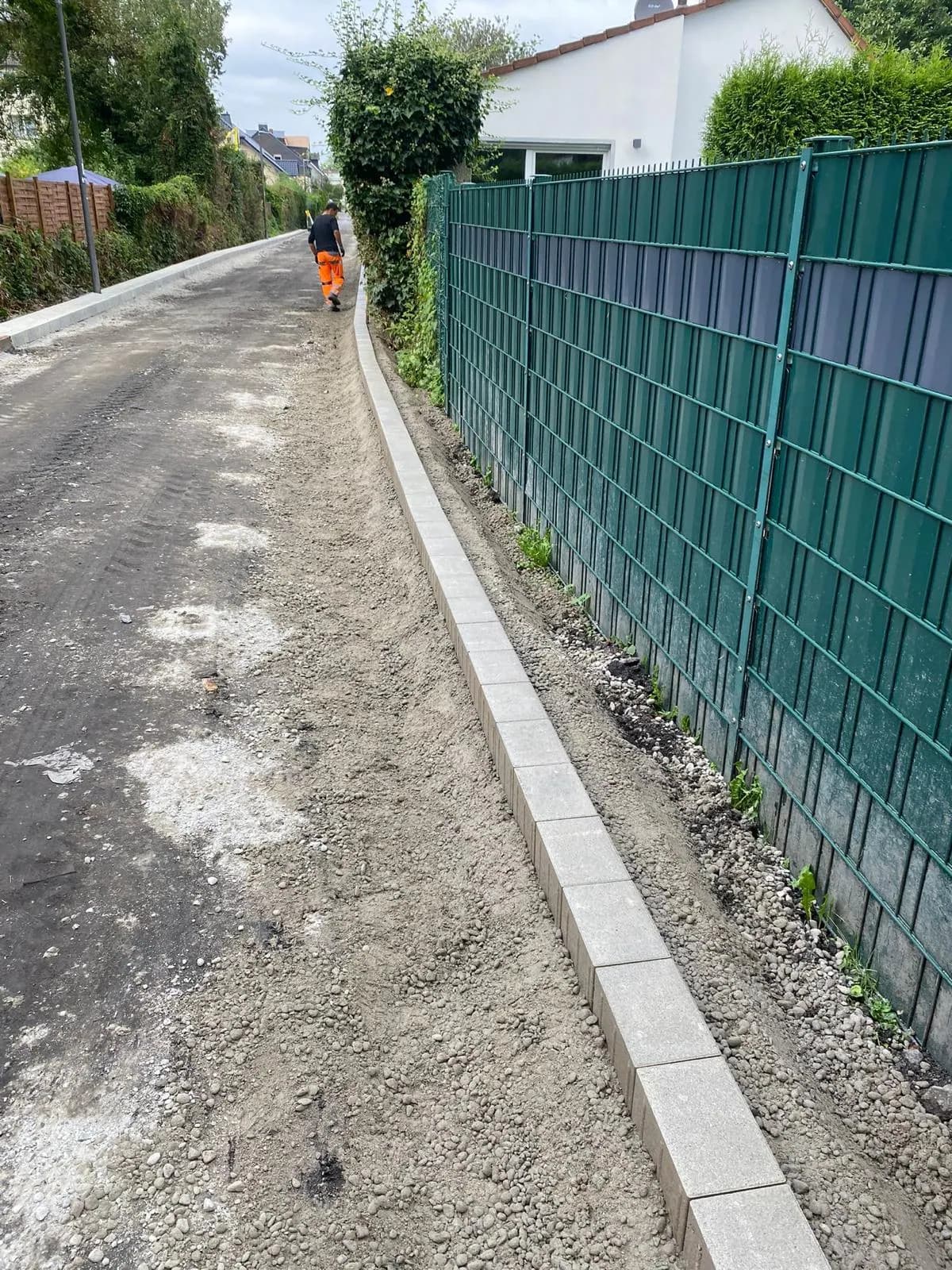 Asphaltweg entlang eines grünen Sichtschutzzauns mit neu gesetzter Bordsteinkante