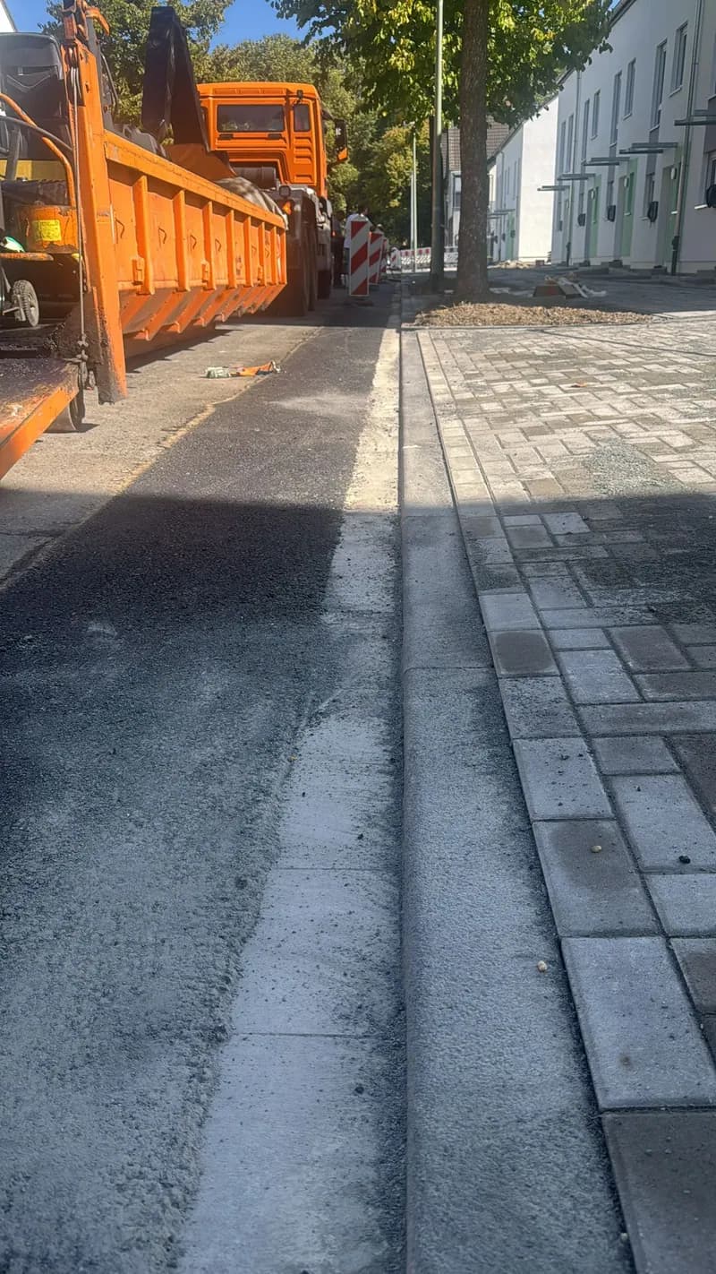 Frisch asphaltierter Fahrbahnrand neben Pflasterstreifen, im Hintergrund ein Container